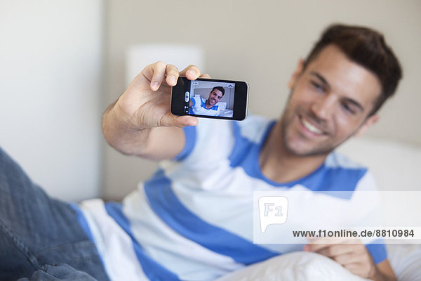 Young man reclining on bed taking selfie with smartphone