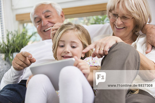 Seniorenpaar und Enkelin sitzend mit digitalem Tablett auf Sofa im Wohnzimmer