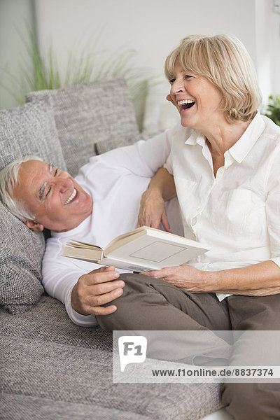 Lachendes Seniorenpaar mit Buch auf Sofa im Wohnzimmer