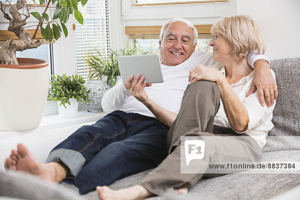 Seniorenpaar mit digitalem Tablett nebeneinander auf Sofa im Wohnzimmer