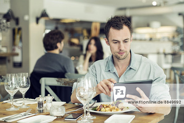Mittlerer erwachsener Geschäftsmann mit digitalem Tablett beim Mittagessen am Tisch im Restaurant