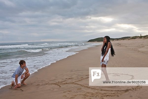 Junger Mann auf einem Knie am Strand  junge Frau in Herzform stehend