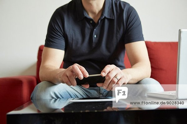 Mittlerer Erwachsener Mann hält Smartphone mit digitalem Tablett und Laptop auf Tisch