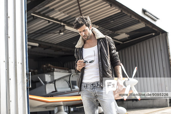 Man with leather jacket standing in front of propeller plane in hangar