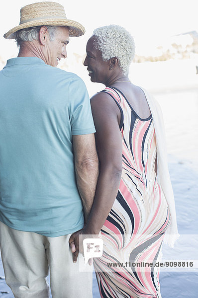 Seniorenpaar hält sich an den Händen und geht am Strand spazieren.