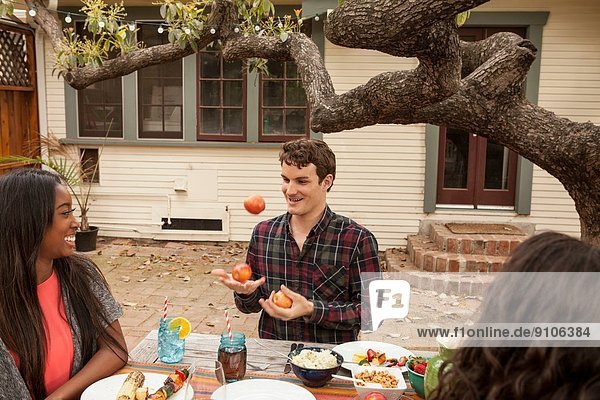 Freunde sitzen am Tisch und teilen sich Grillgut  Mann jongliert mit Früchten