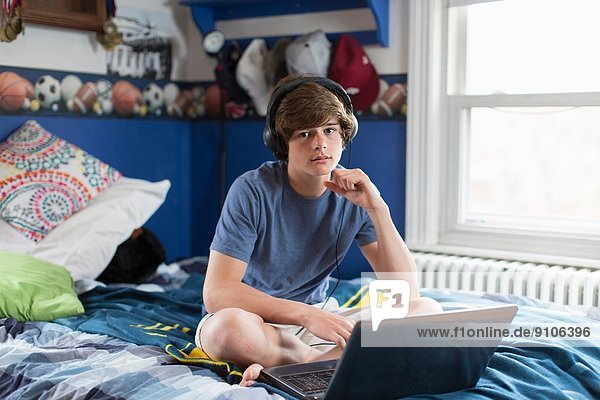 Teenager-Junge auf dem Bett sitzend mit Laptop-Computer