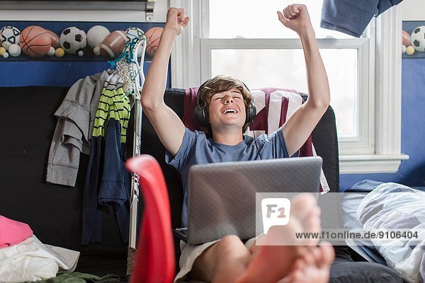 Teenage boy sitting on chair using laptop computer  arms raised