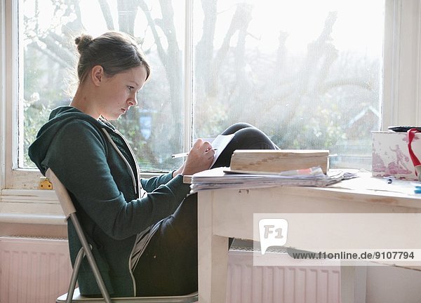 Sechzehnjähriges Mädchen beim Lernen und Schreiben von Notizen neben dem Fenster