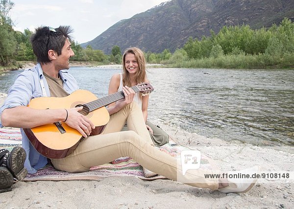 Friends sitting beside river  playing guitar