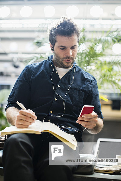 Mittlerer erwachsener Geschäftsmann mit Handy beim Schreiben in einem Buch im Cafe