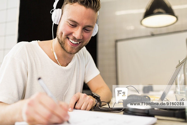 Glücklicher junger Geschäftsmann mit Kopfhörer beim Schreiben im Kreativbüro