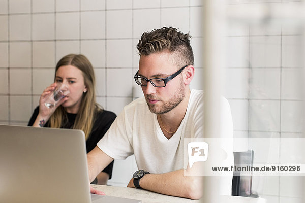 Young businessman working on laptop with female colleague having glass of water in new office