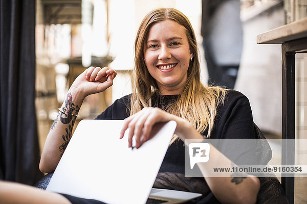 Porträt einer glücklichen jungen Geschäftsfrau mit Laptop auf Bohnentüte im kleinen Büro