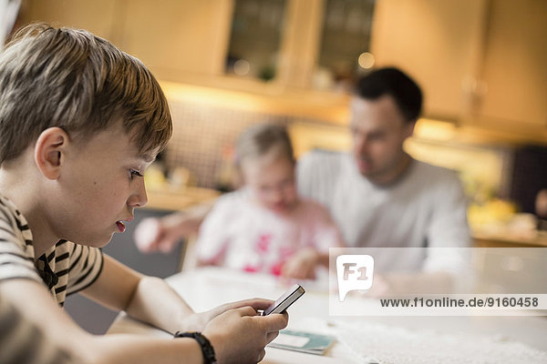 Side view of boy using mobile phone with sister and father in background at home