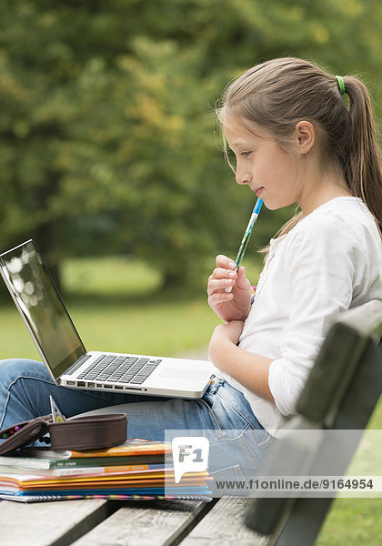 Mächen sitzt auf Parkbank  macht Hausaufgaben mit Laptop