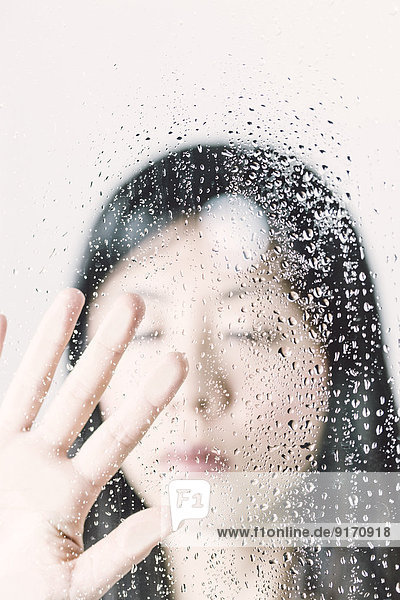 Asiatin mit geschlossenen Augen hinter der Fensterscheibe mit Wassertropfen