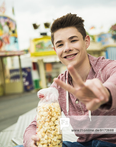 Portrait of happy teenage boy with popcorn showing victory-sign at fun fair