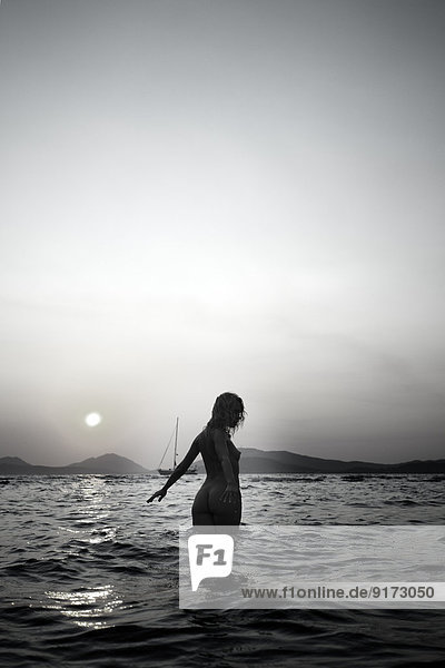 Nackte junge Frau  die in der Dämmerung im Meer spazieren geht.
