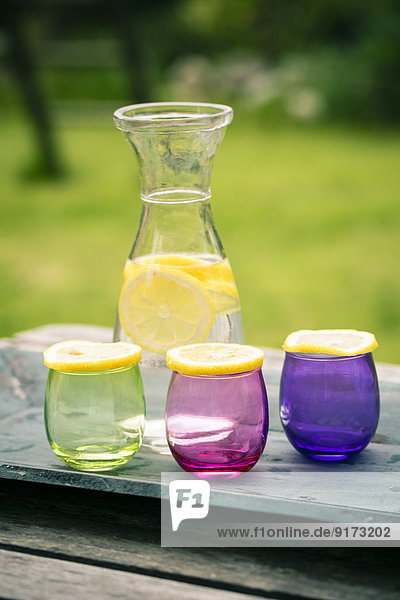 Holztablett  Limonadenkaraffe und Zitronenscheiben auf bunten Gläsern