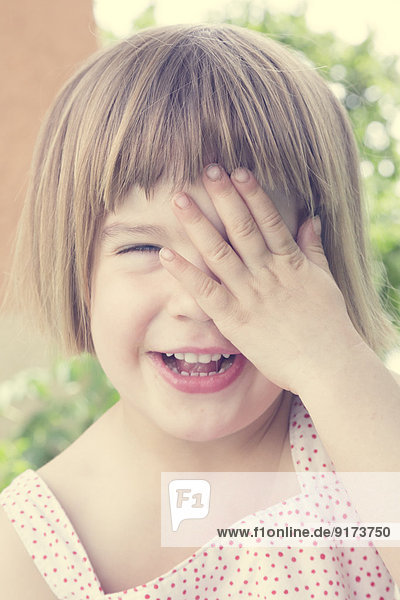 Porträt eines lachenden kleinen Mädchens  das ein Auge mit der Hand bedeckt.