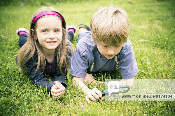 Brother and sister lying on meadow watching flowers with magnifying glass