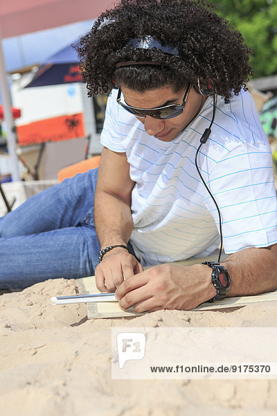 Junger Mann mit digitalem Tablett beim Musikhören am Strand