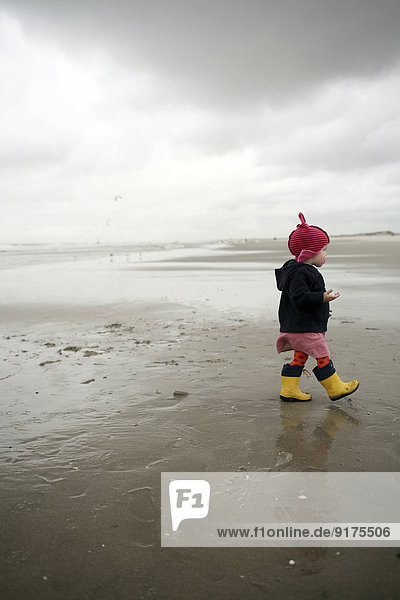 Niederlande,  Rotterdam,  kleines Mädchen mit roter Mütze und gelben Gummistiefeln,  das am nassen Sandstrand spazieren geht