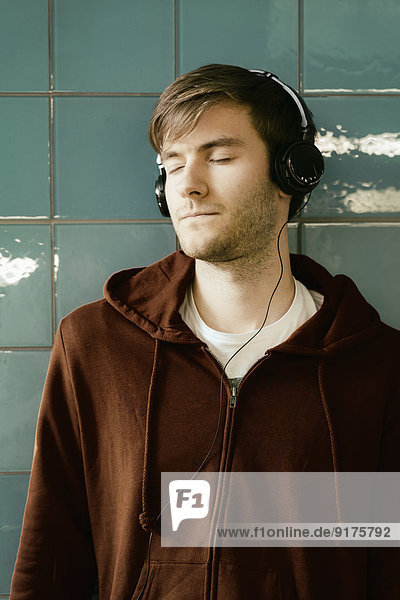 Junger Mann mit Kopfhörer  der sich an eine geflieste Wand lehnt
