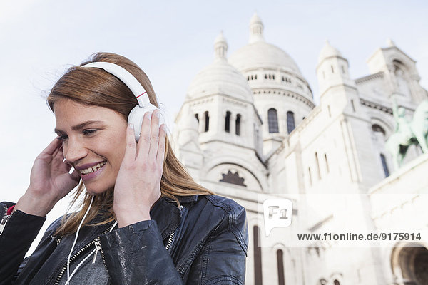 Frankreich  Paris  Porträt einer glücklichen jungen Frau  die Musik mit Kopfhörern vor Sacre Coeur hört.