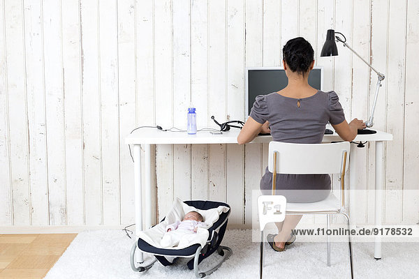 Frau arbeitet im Home-Office  während ihr Baby auf dem Hüpfsitz liegt.