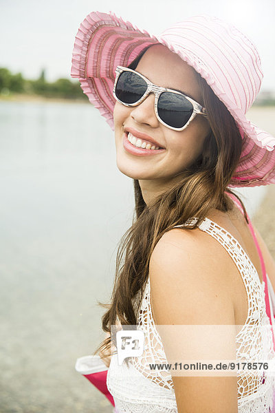 Porträt einer lächelnden jungen Frau mit Sonnenbrille und rosa Hut am Strand