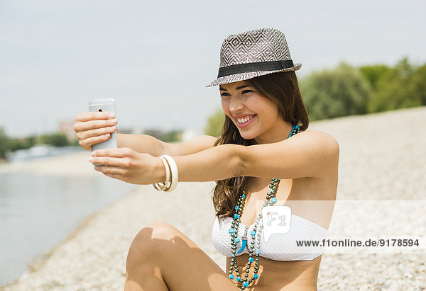 Porträt einer lächelnden jungen Frau  die einen Selfie mit ihrem Smartphone am Strand nimmt.