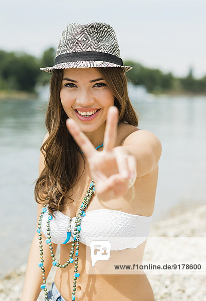 Porträt einer lächelnden jungen Frau mit Siegeszeichen am Strand