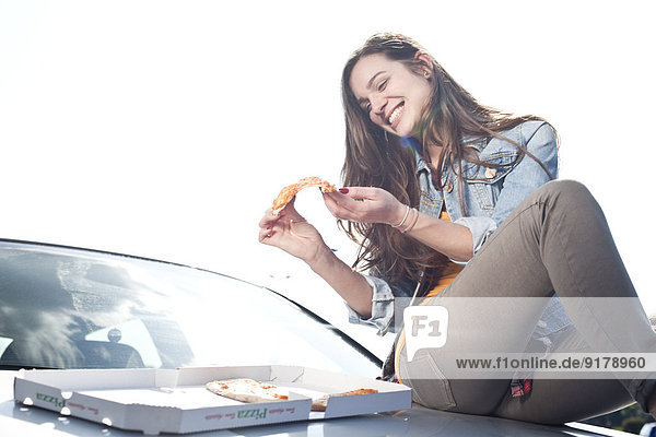 Fröhliche junge Frau beim Pizzaessen auf der Autohaube