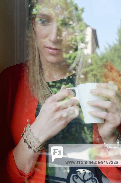 Junge Frau mit Kaffeetasse aus dem Fenster blickend