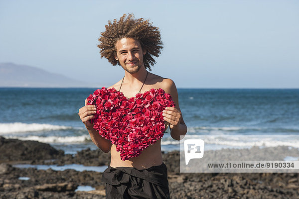 Porträt eines lächelnden jungen Mannes mit herzförmigen Blumen am Strand