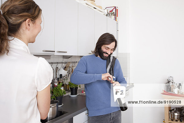 Woman looking at smiling man opening wine bottle in kitchen