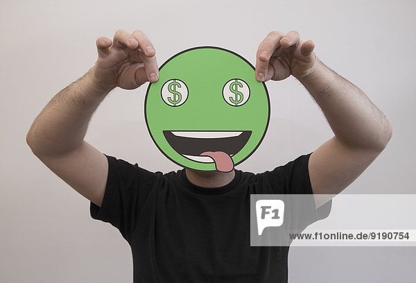 Mann hält ein grünes Dollarzeichen Emoticon-Gesicht vor seinem Gesicht.