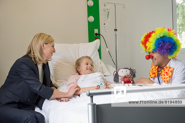 Male doctor wearing clown costume making girl patient smile in hospital bed