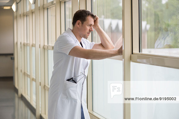 Männlicher Arzt steht im Krankenhaus und schaut aus dem Fenster.