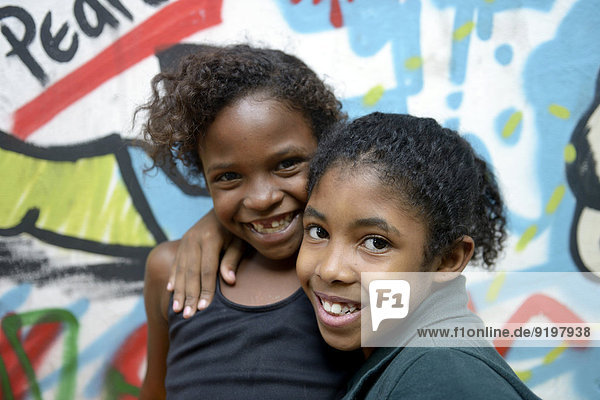 Two smiling girls embracing each other in front of a graffiti-sprayed wall  slum  Mangueirinha favela  Duque de Caxias  Rio de Janeiro  Brazil