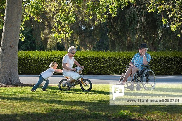 Three year old boy pushing grandmother on cycle with grandfather watching from wheelchair