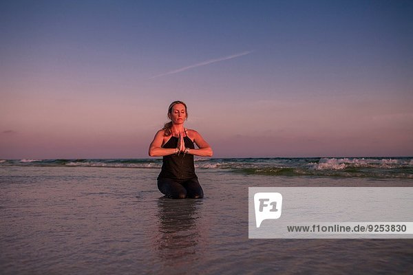 Reife Frau  die am Strand bei Sonnenuntergang kniend Yoga praktiziert.