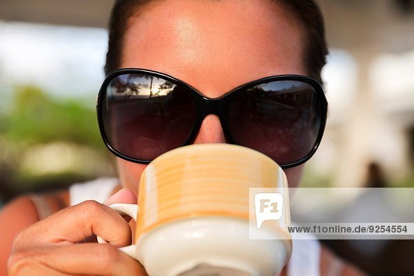 Nahaufnahme einer erwachsenen Frau in einer Sonnenbrille  die aus einer Kaffeetasse trinkt.