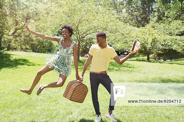 Junges Paar mit Picknickkorb im Park  mit weiblichem Sprung in der Luft