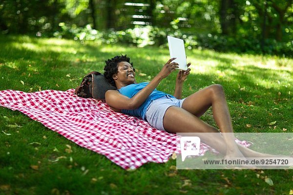 Junge Frau liegt im Park und schaut auf das digitale Tablett.