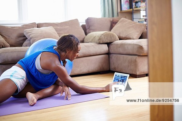 Junge Frau  die auf dem Boden des Wohnzimmers trainiert  während sie den Touchscreen auf dem digitalen Tablett benutzt.