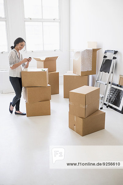 Young woman standing among boxes in new home and using mobile phone