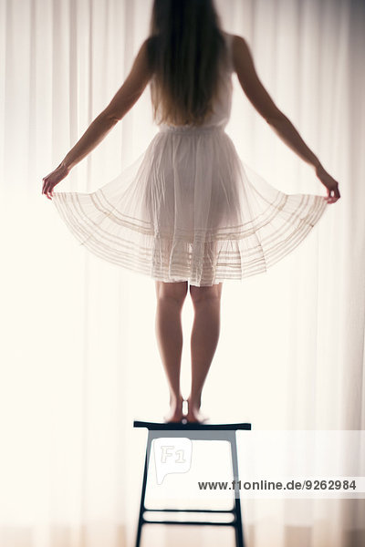 Junge Frau steht auf einem Hocker und hält die Naht ihres Rockes vor einem weißen Vorhang  Rückansicht